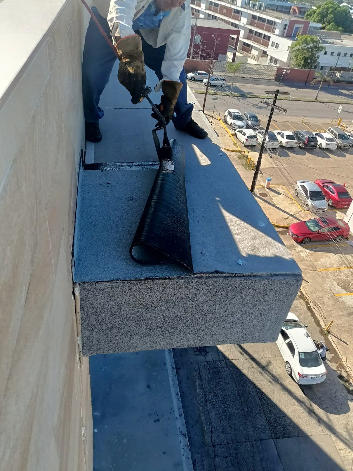 Técnico aplicando membrana en pretil a altura, estacionamiento visible abajo